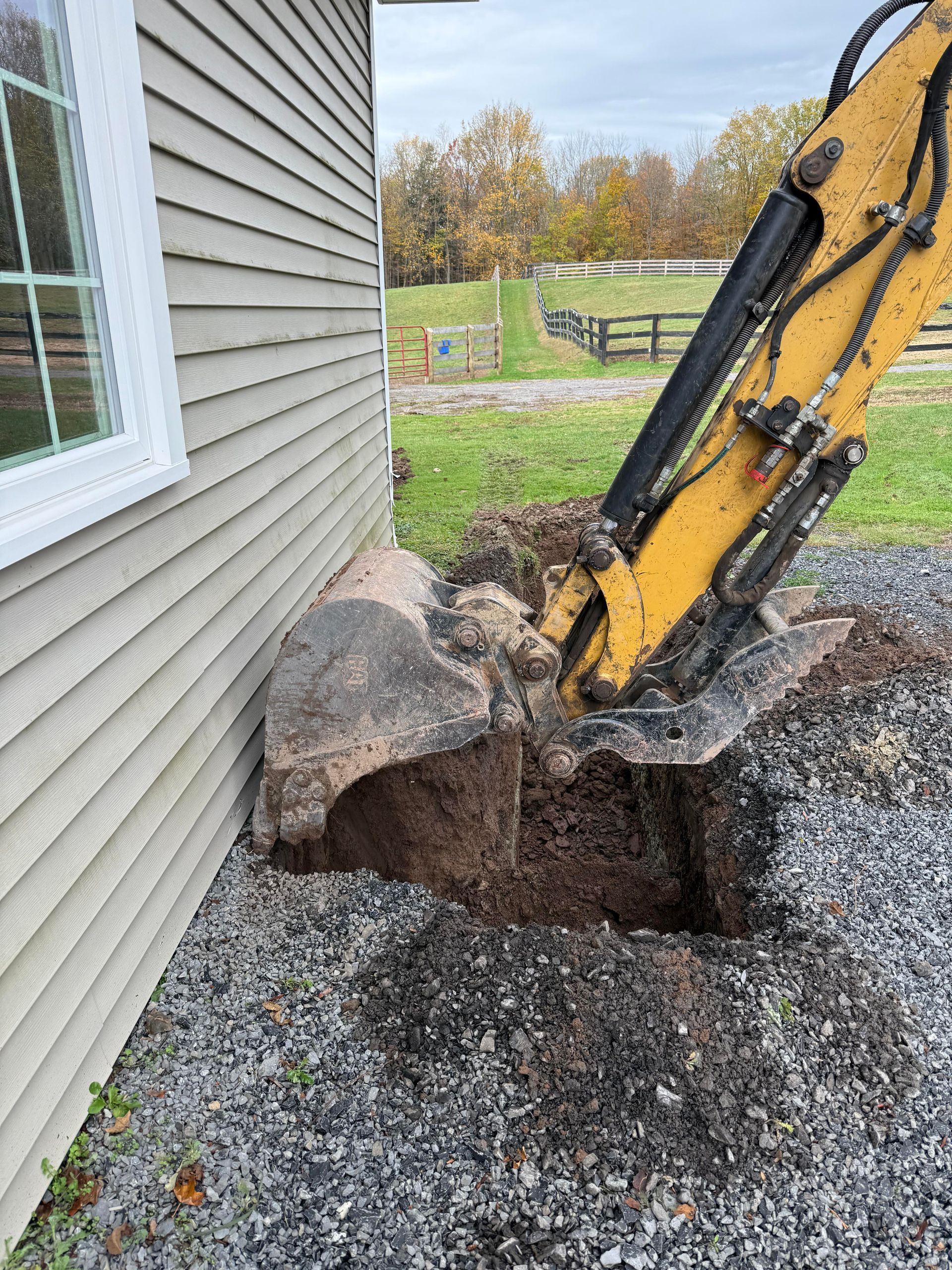 Yellow excavator digging near a building's gray siding, creating a hole in gravel.