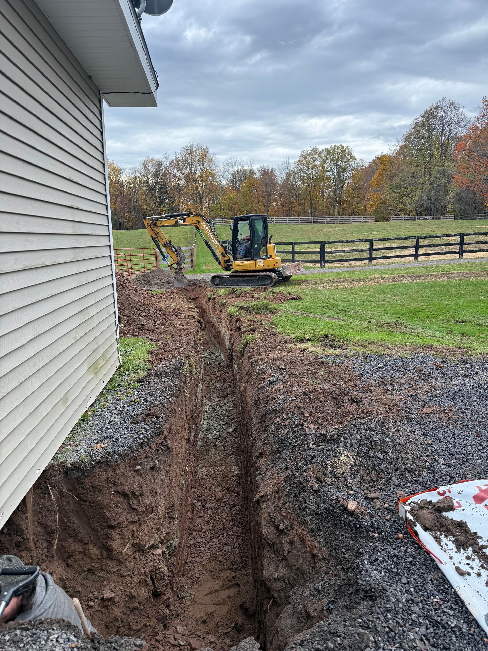 A small excavator digs a trench next to a house; the sky is overcast, and trees are in the background.