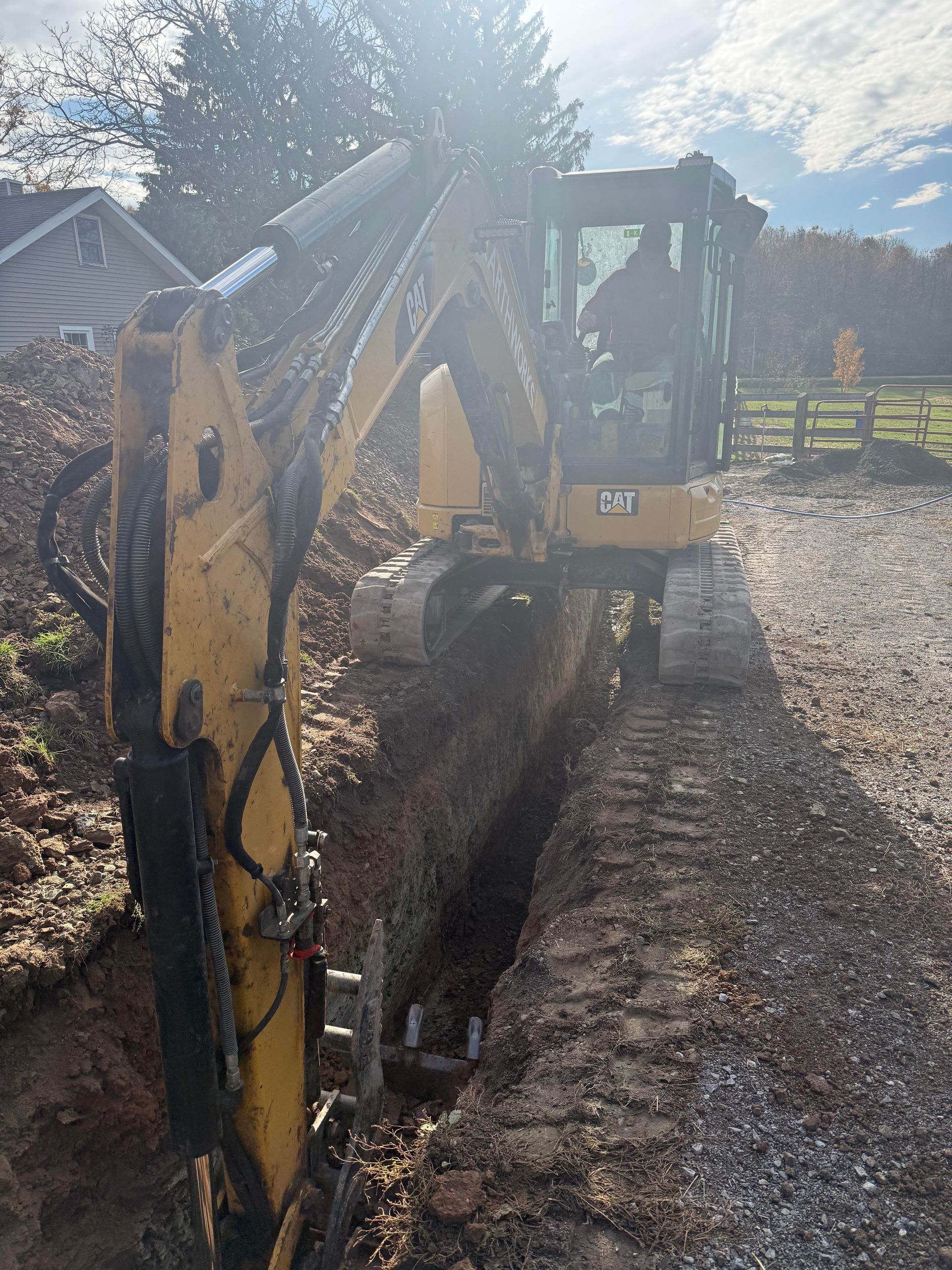 Yellow Caterpillar excavator digging a trench in a gravel driveway; a person operates the machine.