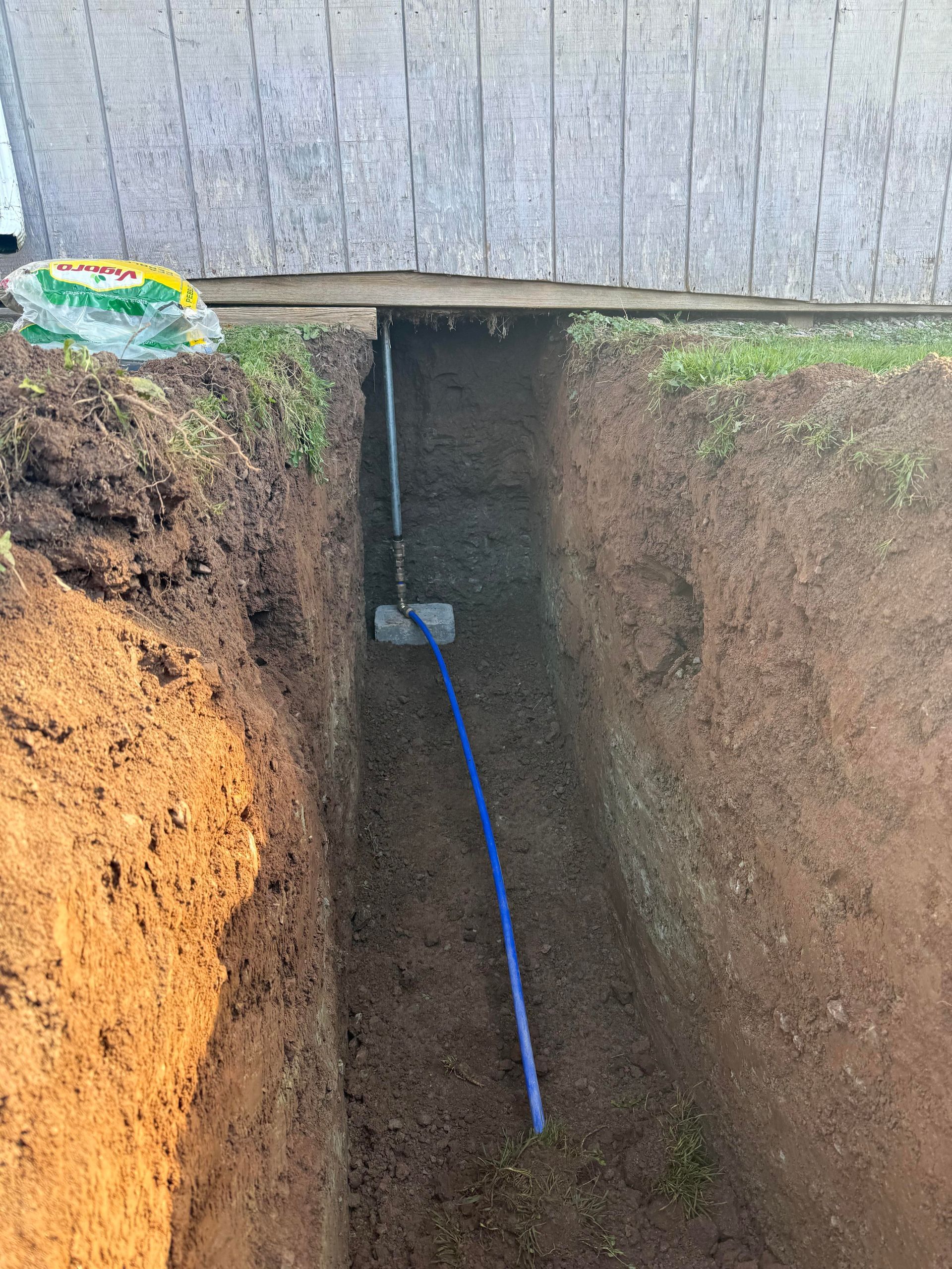 Trench dug near a building. Blue pipe runs down the trench, possibly for utilities.