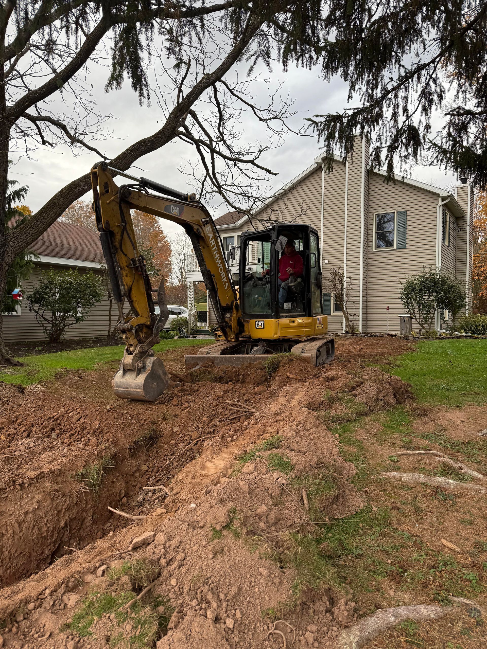 Yellow excavator digging a trench in a grassy yard, near a light-colored house. Person in the cab.
