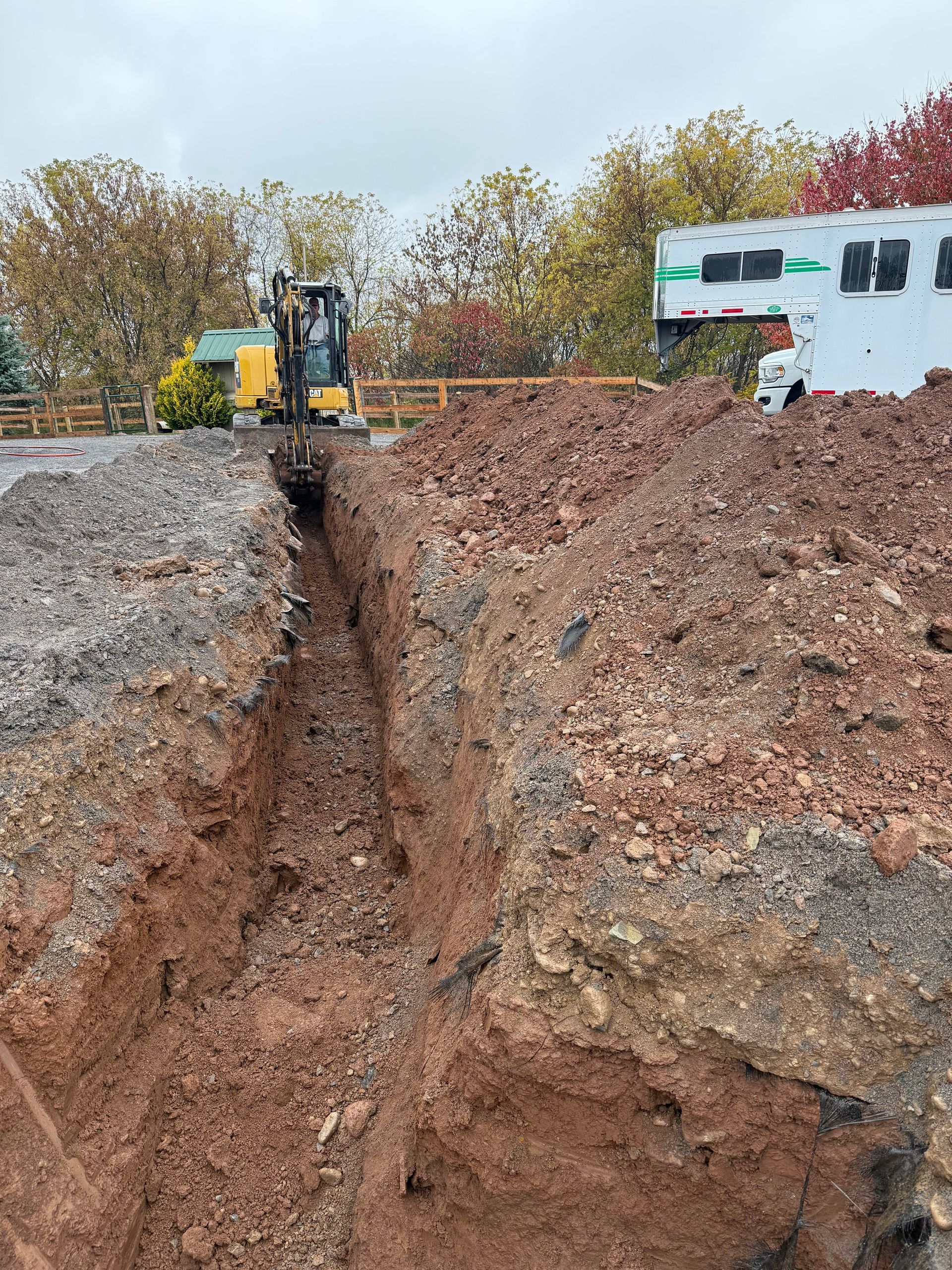 An excavator digs a long trench in reddish-brown soil, a building visible in the background.