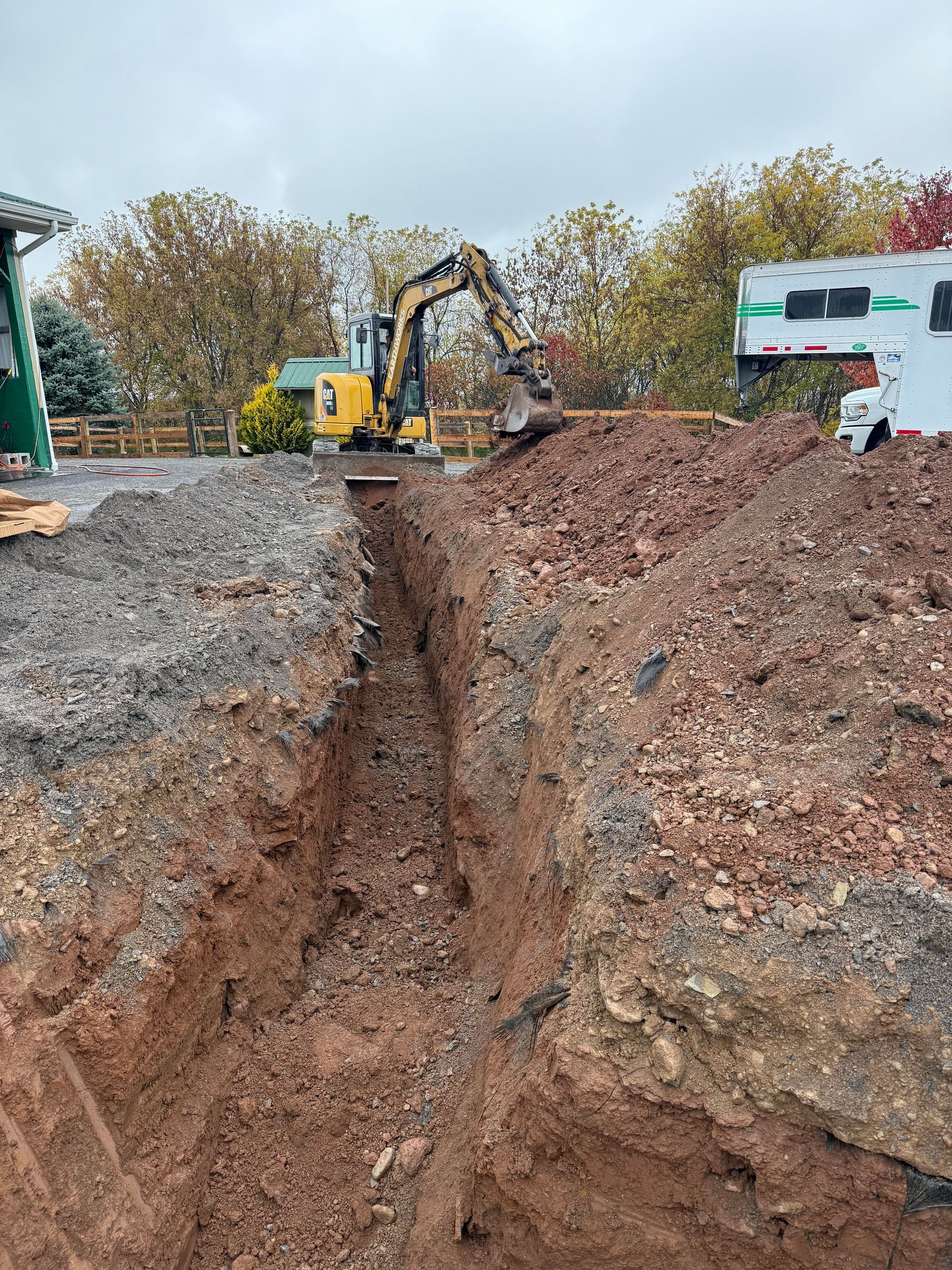 Excavator digging a trench in dirt, near a building and trees. Overcast sky.