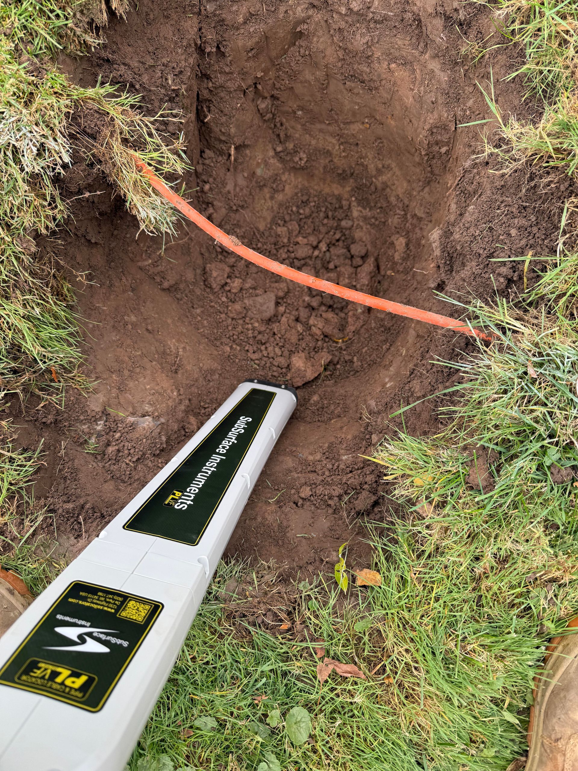 Hole in ground with orange cable. Utility locator in frame. Green grass setting.