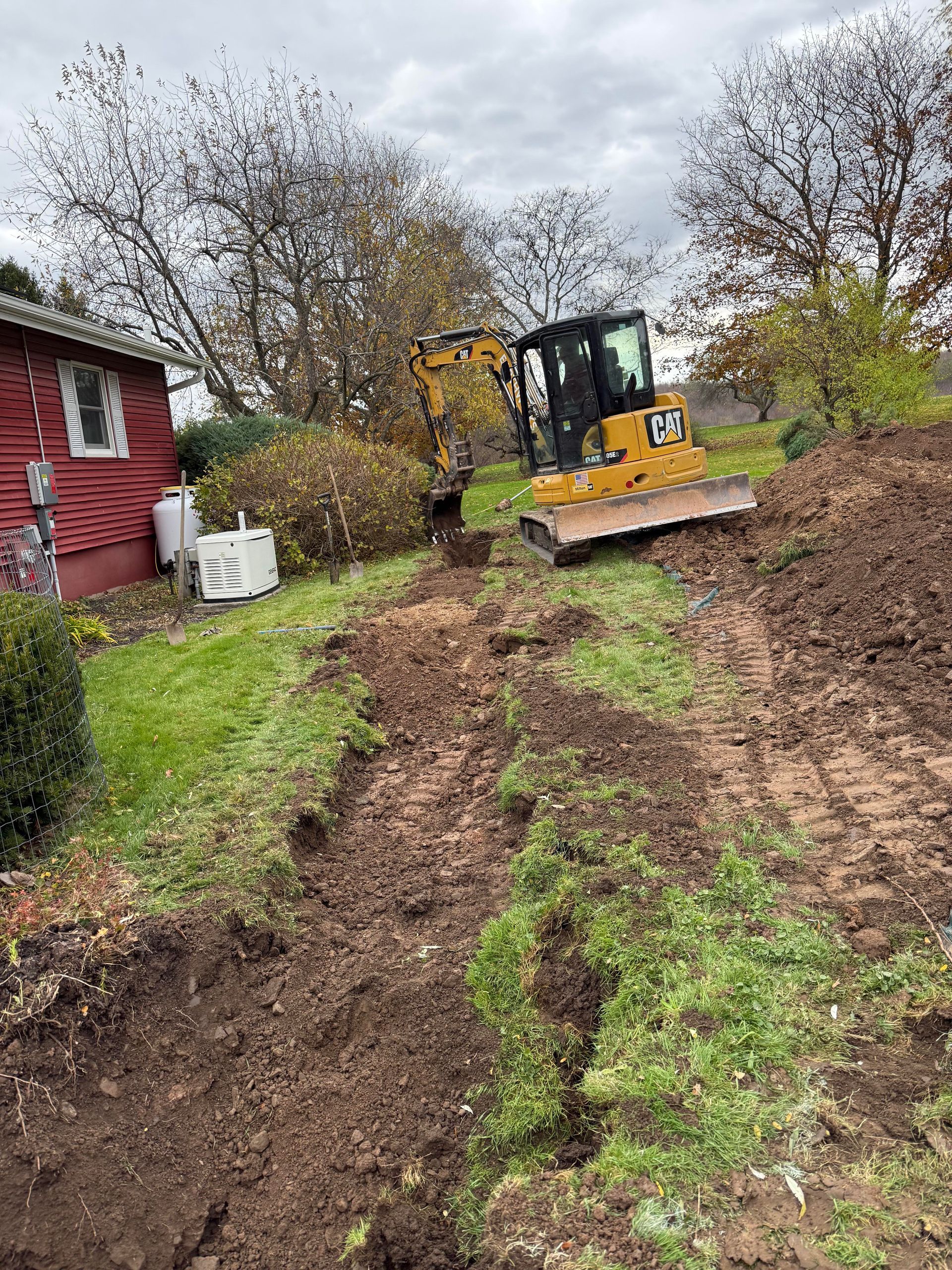 A yellow construction excavator digs a trench in a grassy yard next to a red building on a cloudy day.