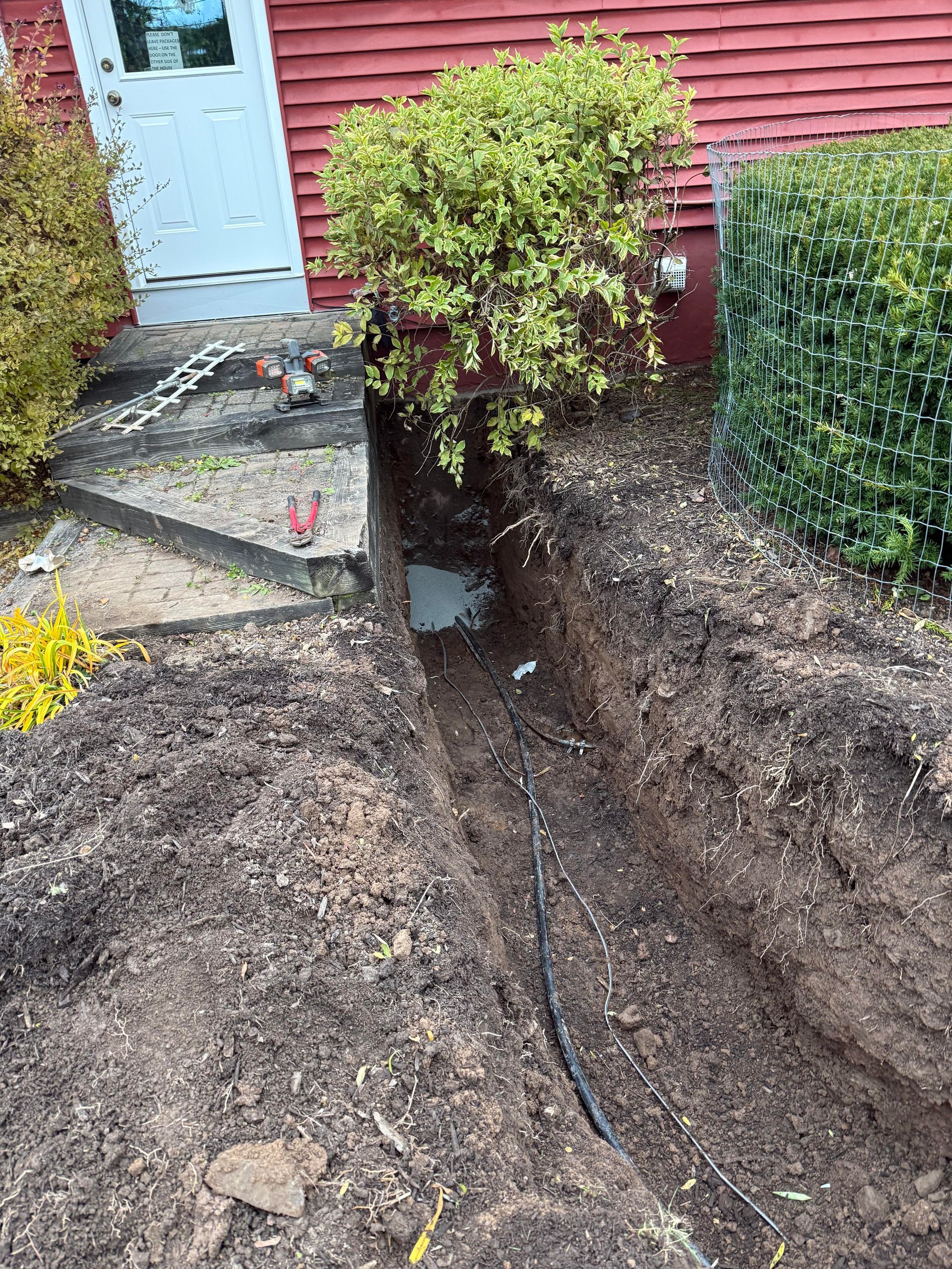 Trench dug in front of a red building, exposing a black cable. Soil and green shrubs surround the trench.