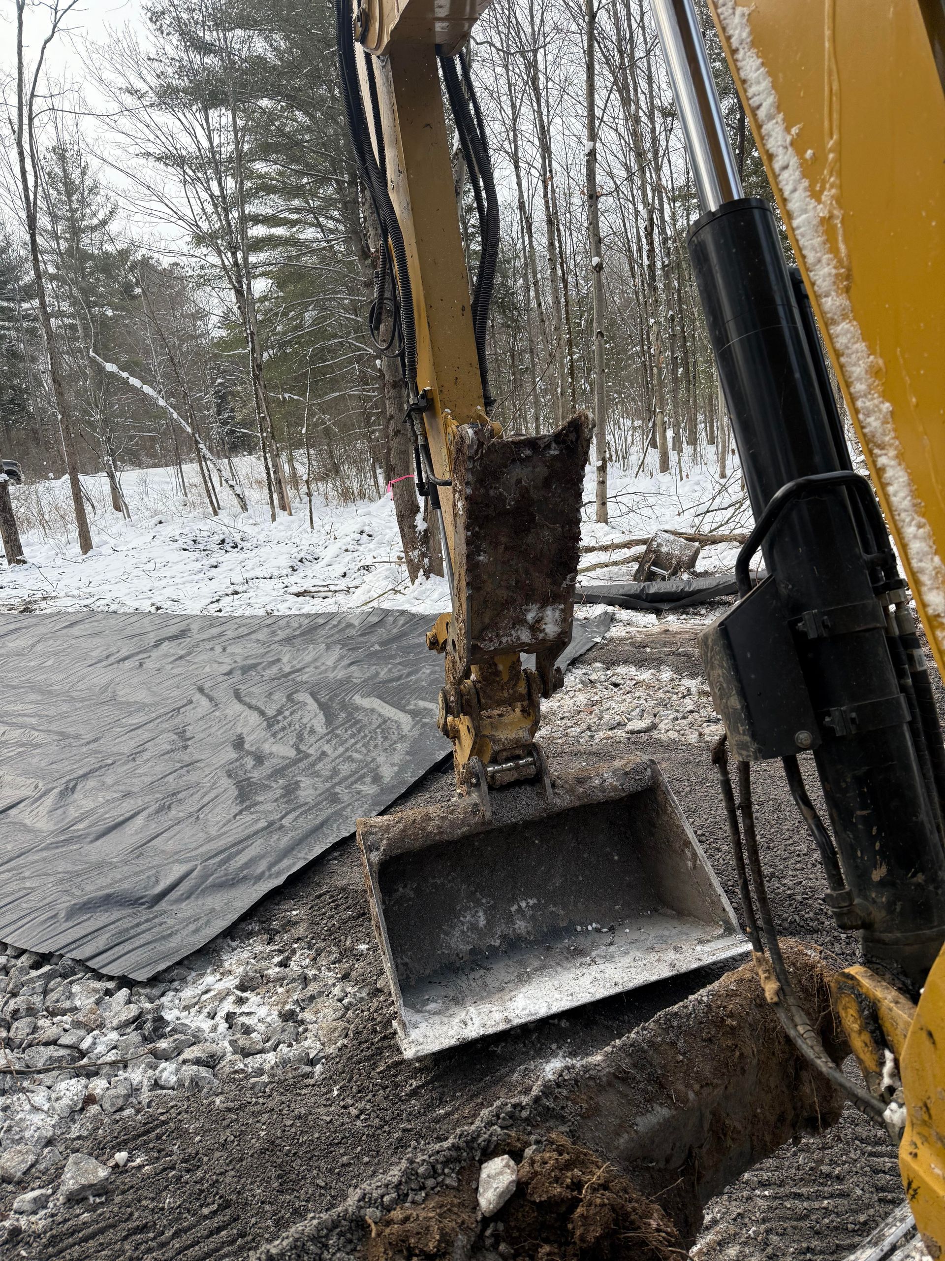 Yellow excavator arm and bucket in snow, digging into the earth. Black tarp is visible.