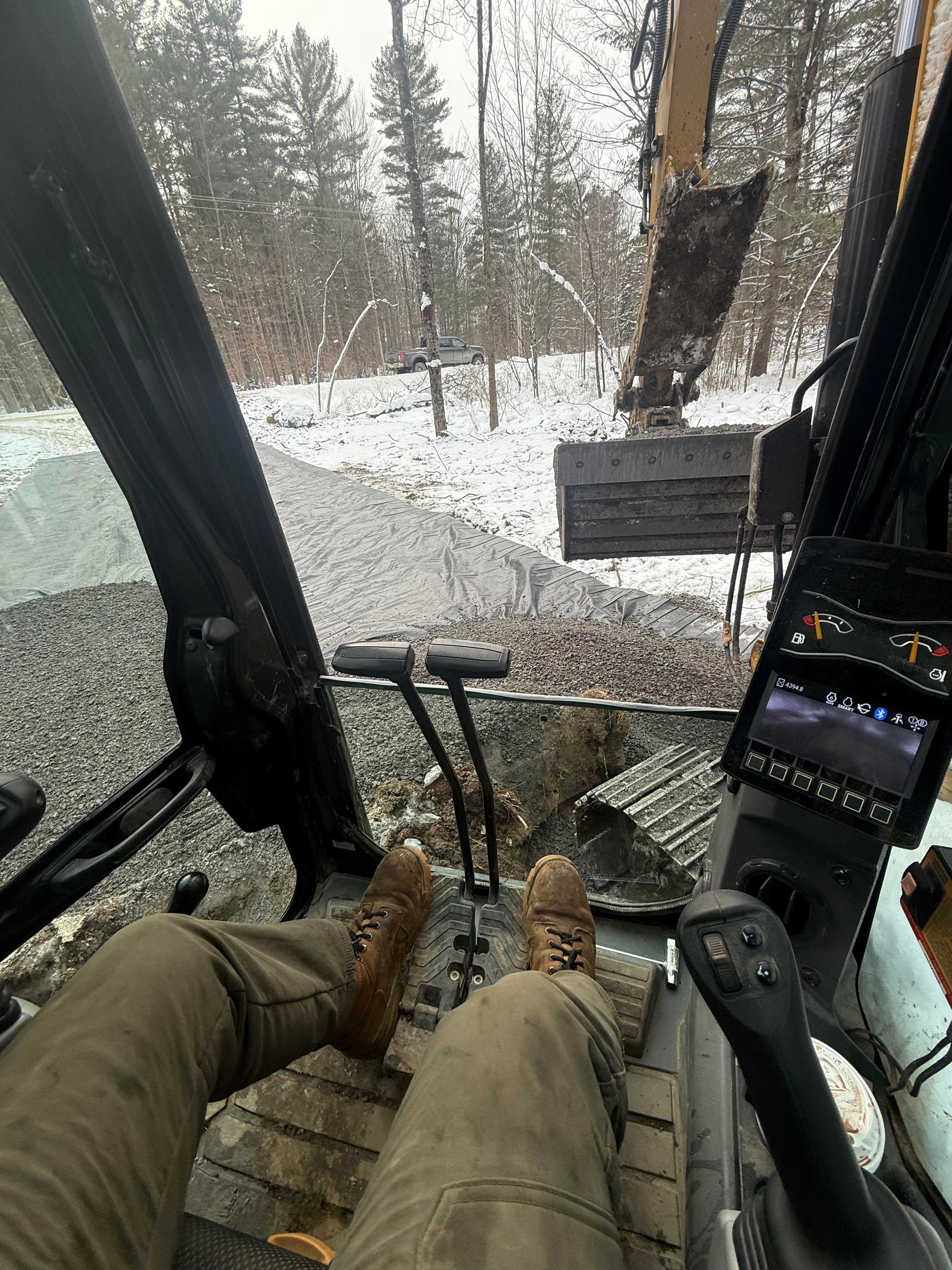 View from inside excavator cab, operating controls, legs visible, snowy landscape.