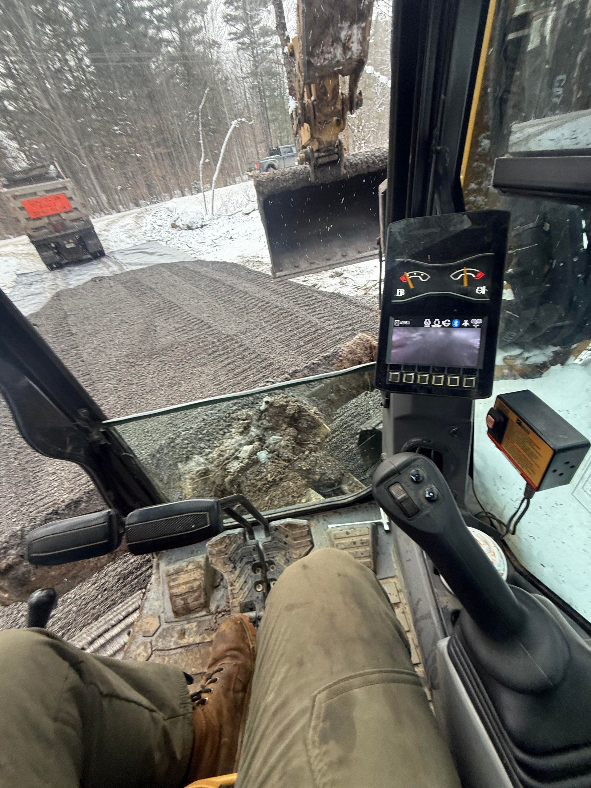 Inside excavator cab, view of operator's legs, controls, and screen showing camera views. Gravel pile and dump truck outside.