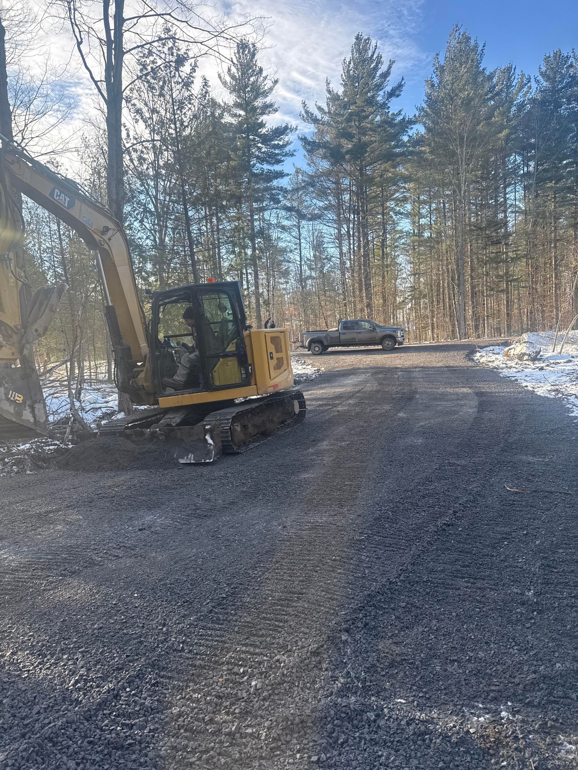 An excavator working on a gravel road, with a pickup truck in the background, set in a wooded area with some snow.