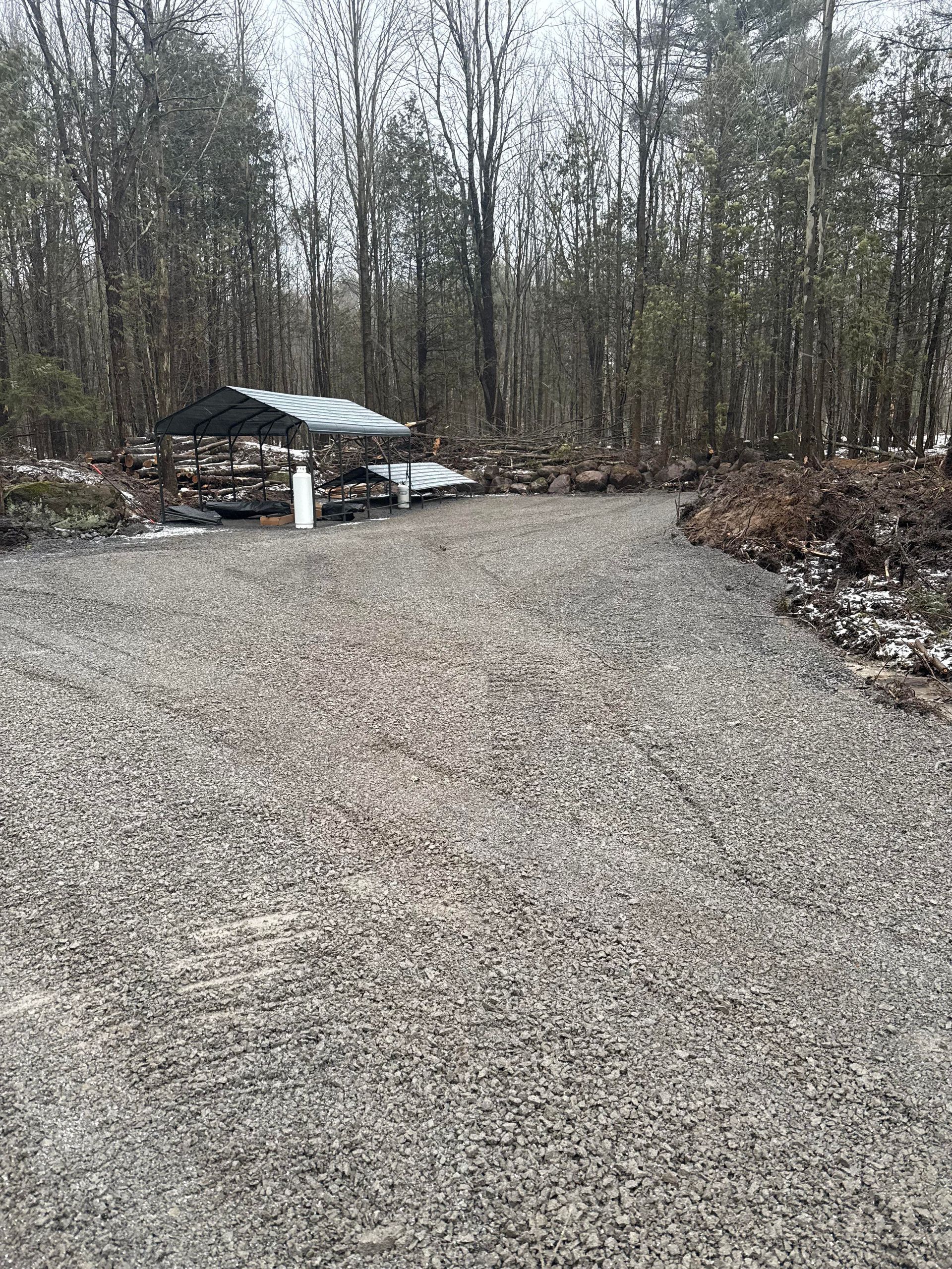 Gravel driveway leading to a metal-roofed shelter in a wooded area. Grey tones dominate, some snow present.