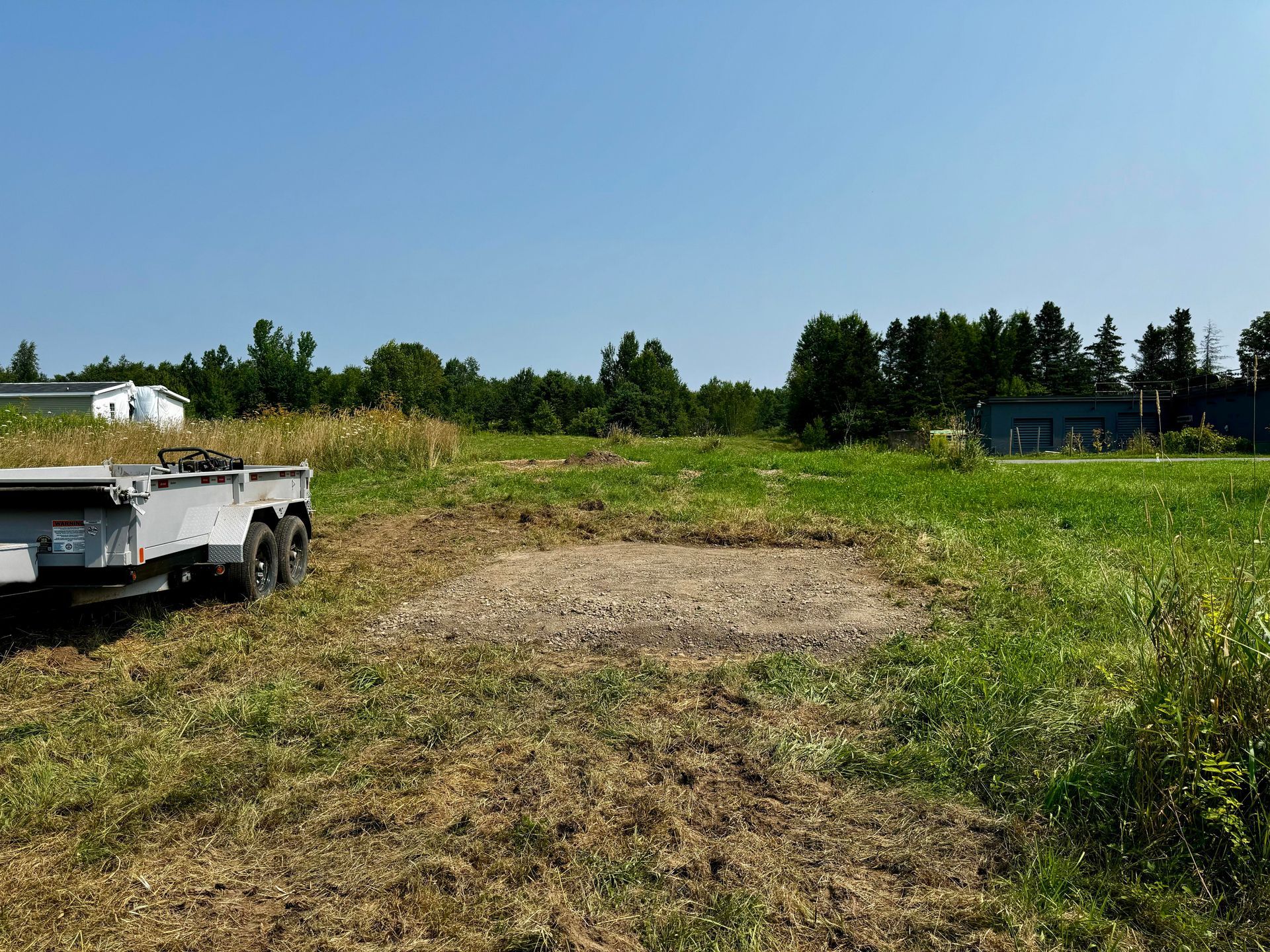 A trailer sits in a grassy field with a gravel patch. Trees and a blue sky are in the background.