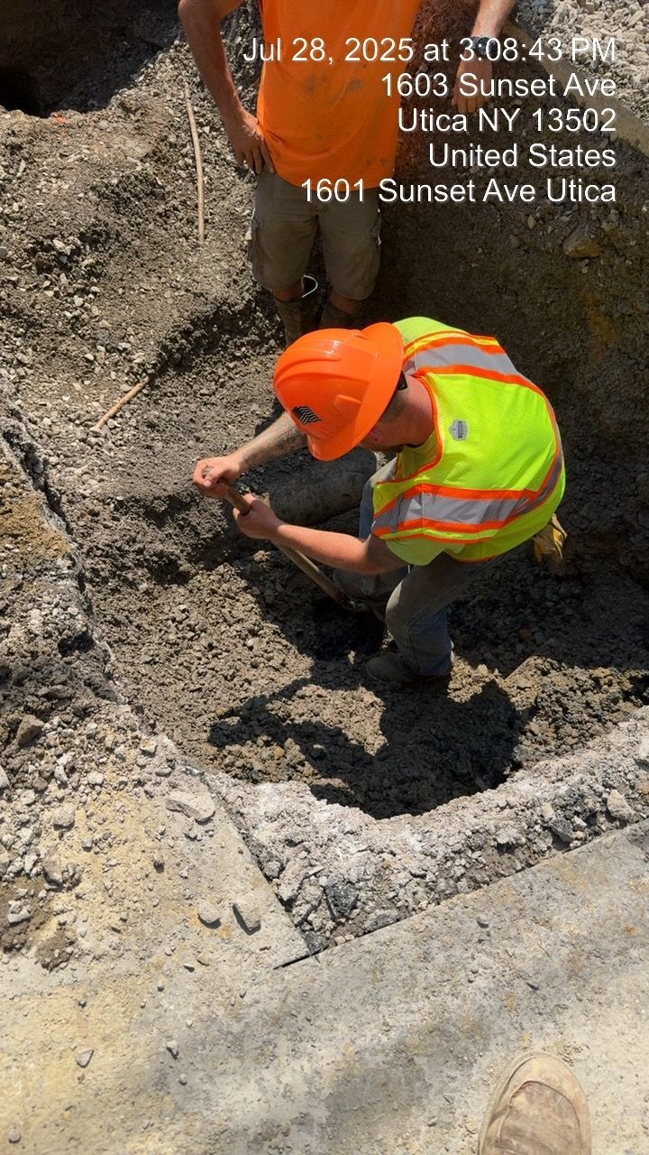 Workers inspecting a ditch. One kneels, wearing a hard hat and safety vest. Another stands nearby.