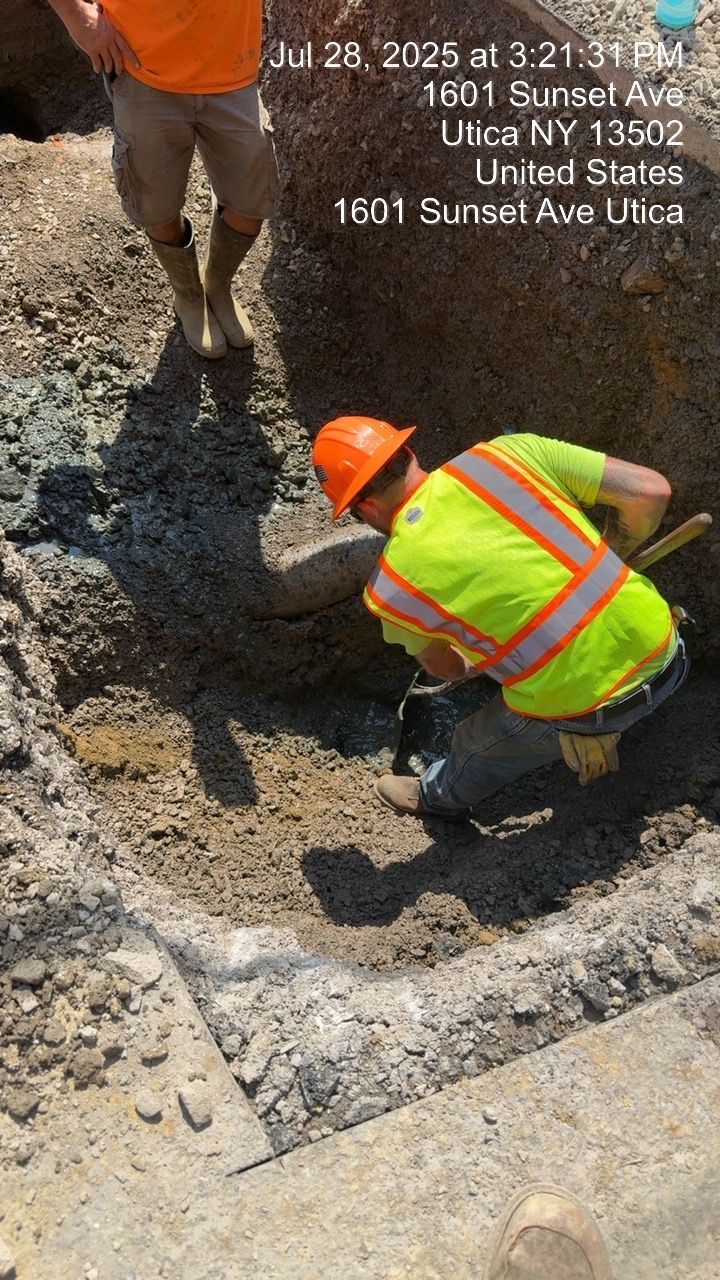 Construction worker digging in a trench at 1601 Sunset Ave, Utica NY. Another person stands nearby. Date and time visible.