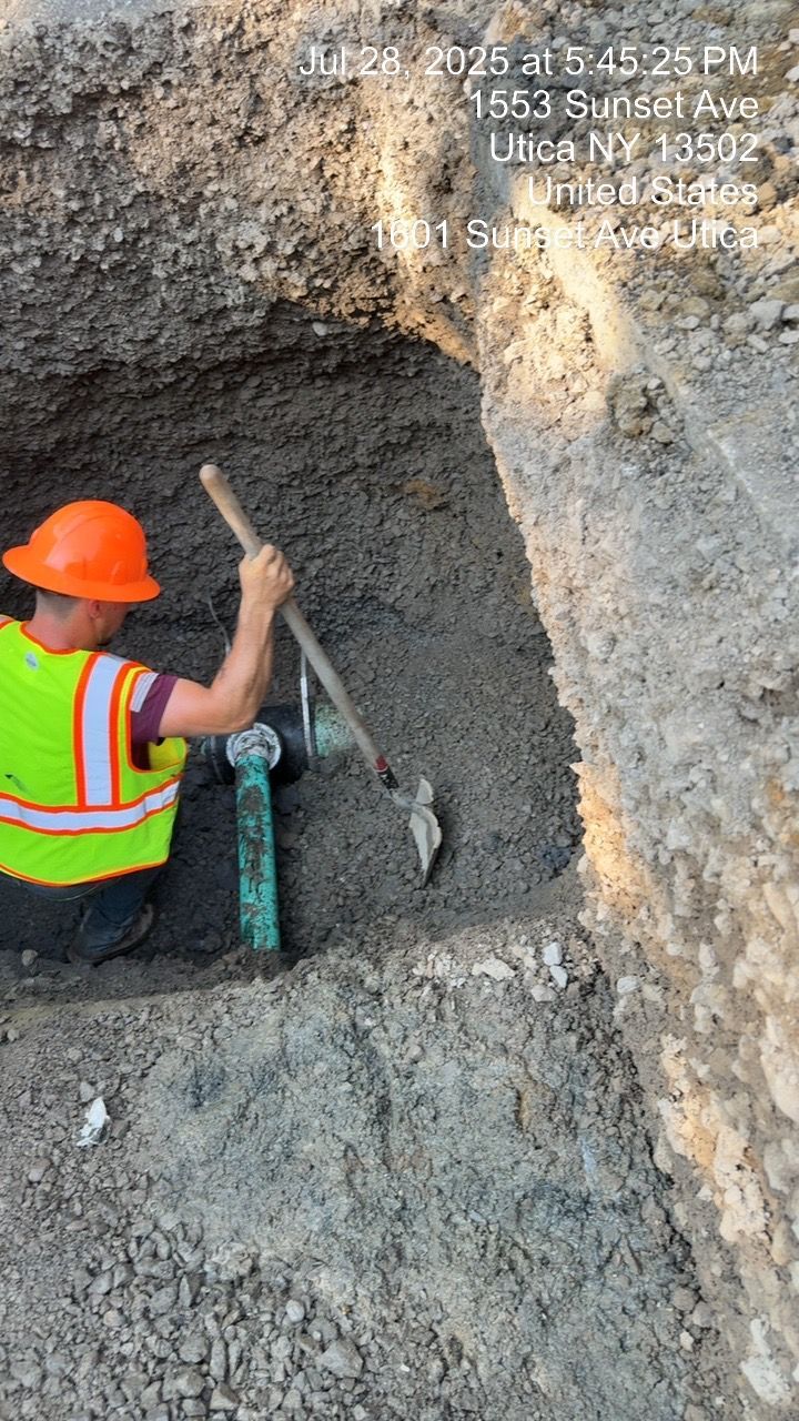 Worker excavating near a green pipe in a ditch, wearing orange hat and reflective vest.