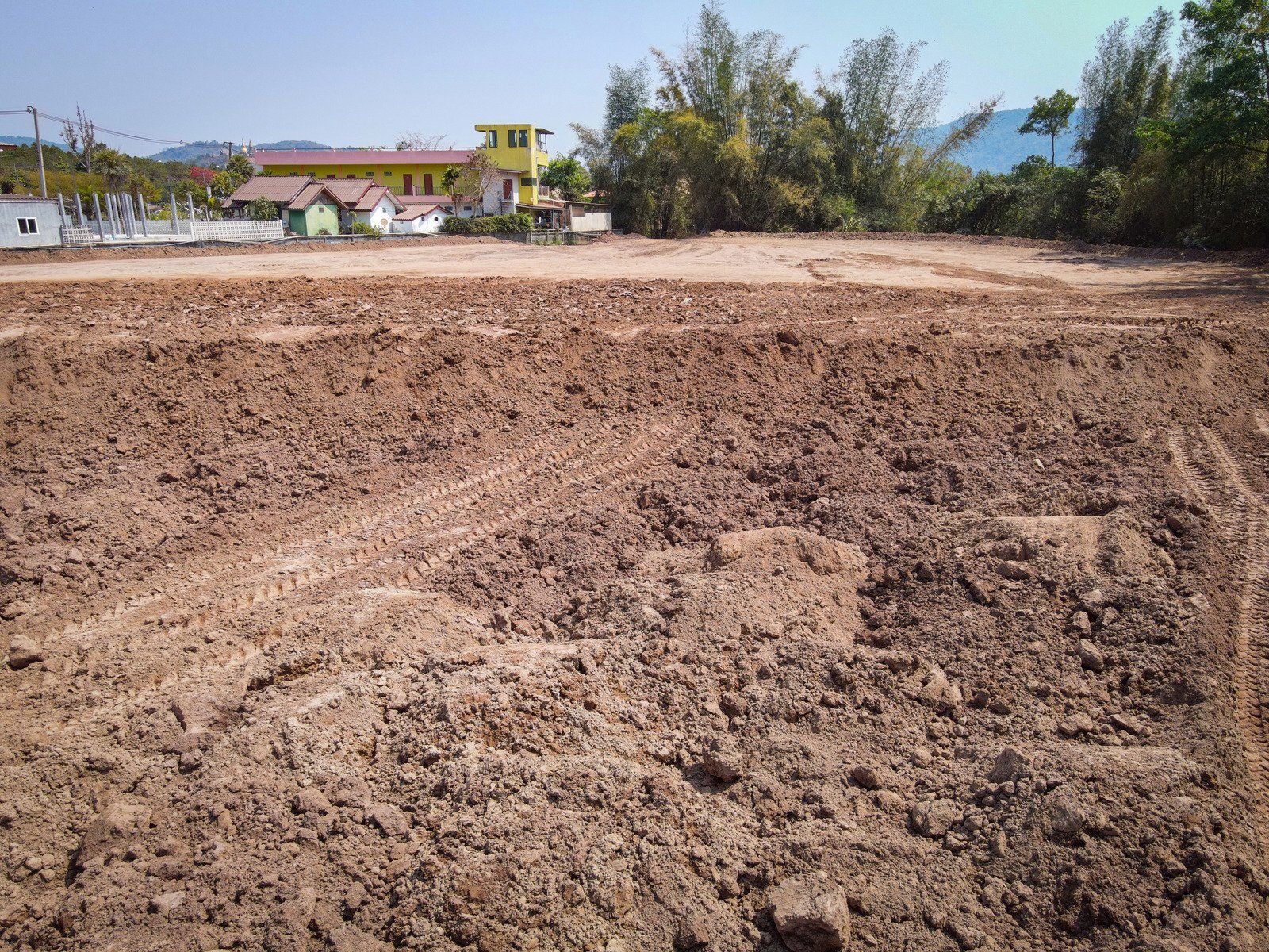 Brown, freshly tilled soil in a large, cleared field, with buildings and trees visible in the background.