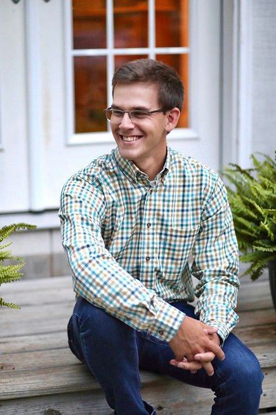A young man wearing glasses and a plaid shirt is sitting on a porch.