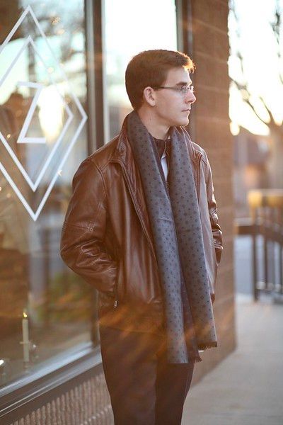 A young man wearing a leather jacket and scarf is standing in front of a store window.
