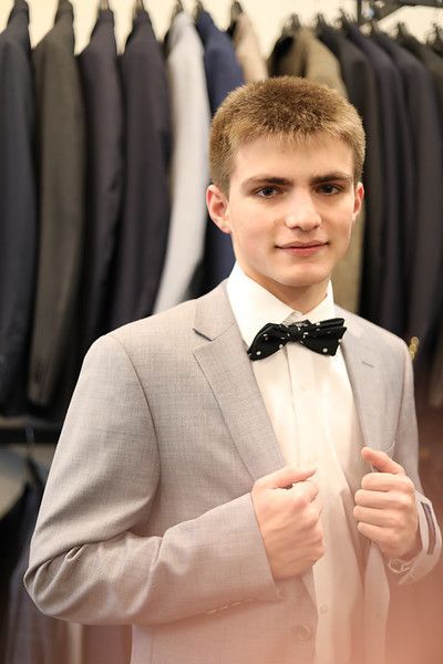 A young man in a suit and bow tie is standing in front of a rack of suits.