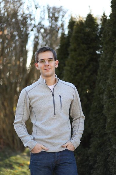 A man in a grey sweater and blue jeans is standing in front of trees.