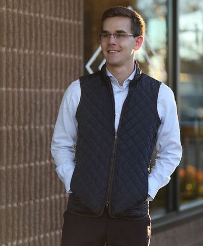 A man wearing a quilted vest and glasses is standing in front of a building.