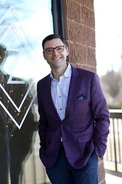 A man in a purple suit and glasses is standing in front of a building.