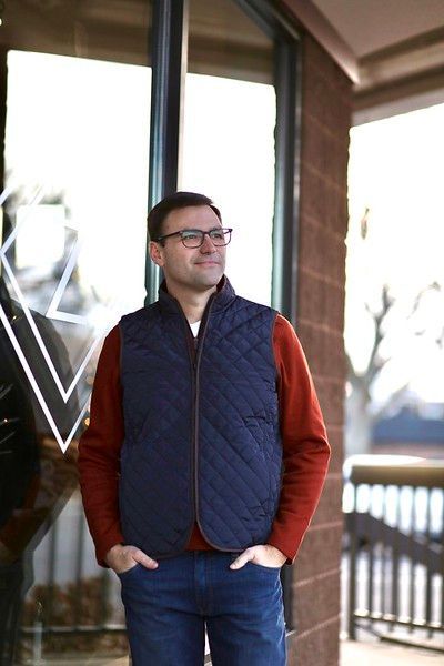 A man wearing a vest and glasses is standing in front of a window.