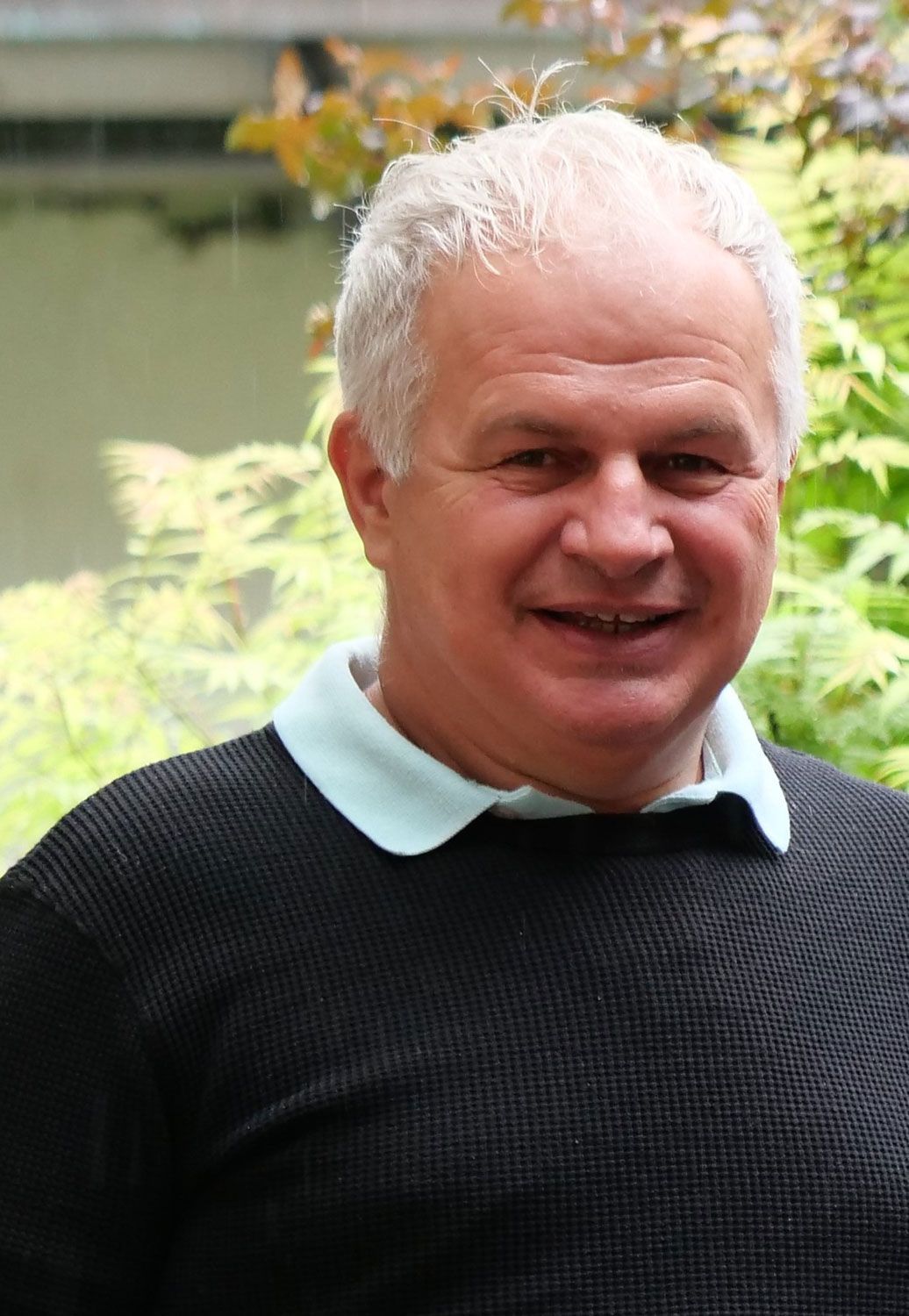 Ein Mann mit weißem Haar trägt einen schwarzen Pullover und einen weißen Kragen.