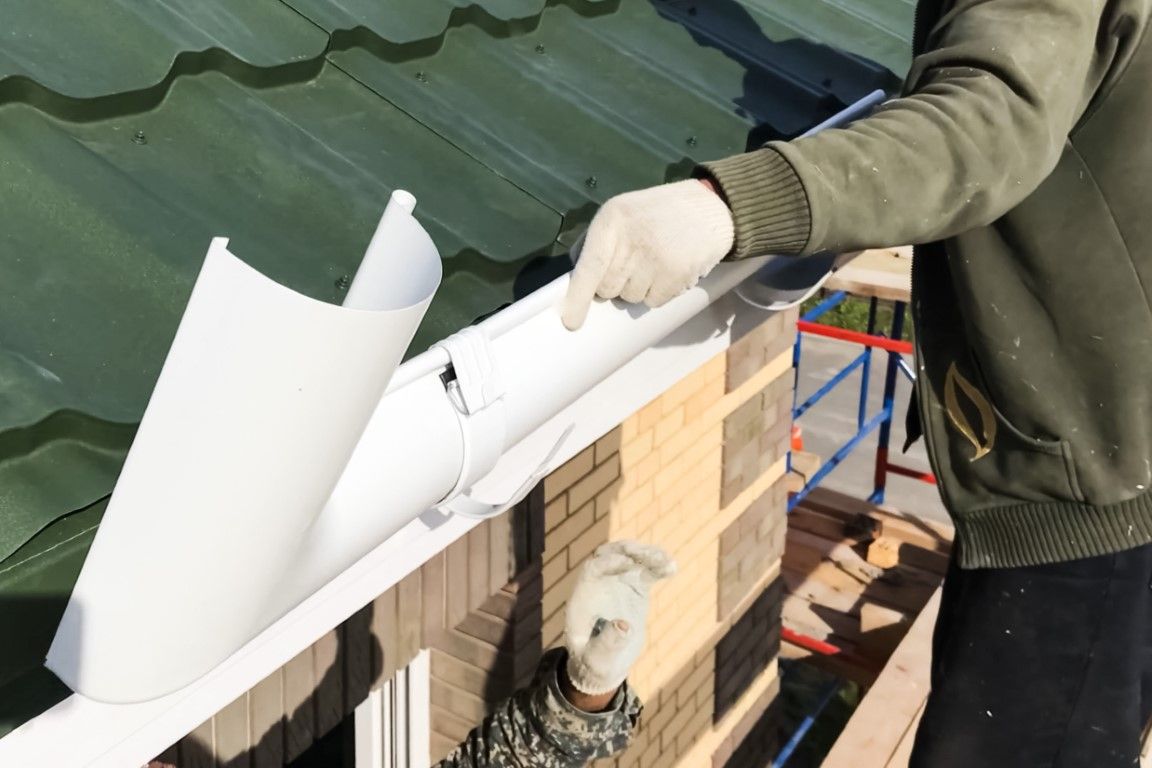 Person installing a white gutter on a brick building with a green roof.