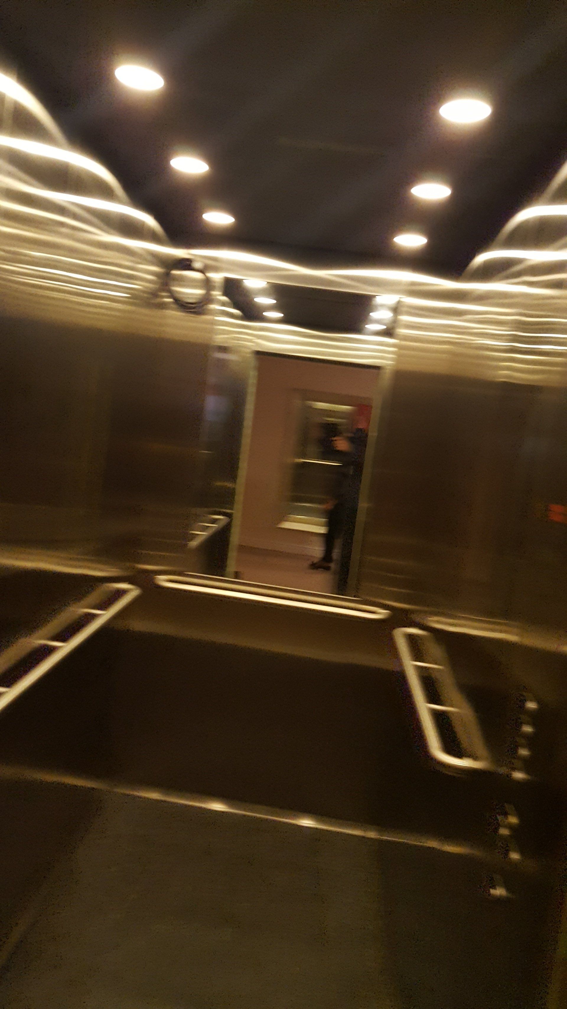 Interior of an elevator with stainless steel walls and ceiling lights. A person stands near open doors.
