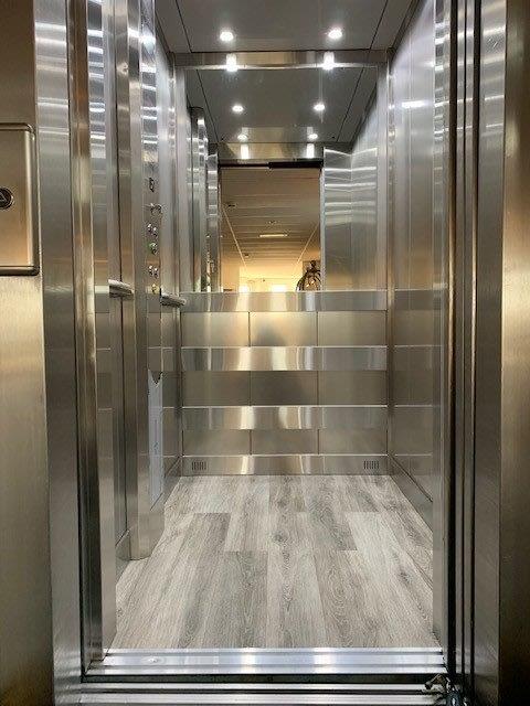 Modern elevator interior with stainless steel walls and wood-look flooring. Doors open, revealing controls.
