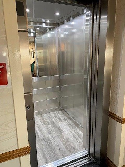 Interior view of an elevator with stainless steel walls, open door, and wood-look floor.