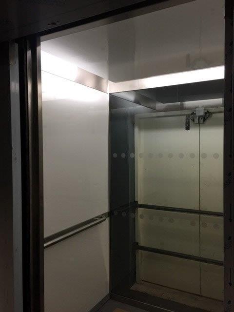 Empty elevator interior with stainless steel and white walls, handrail, and closed glass doors.