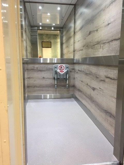 Elevator interior with light gray flooring, wood-look walls, and a mirror reflecting the ceiling.