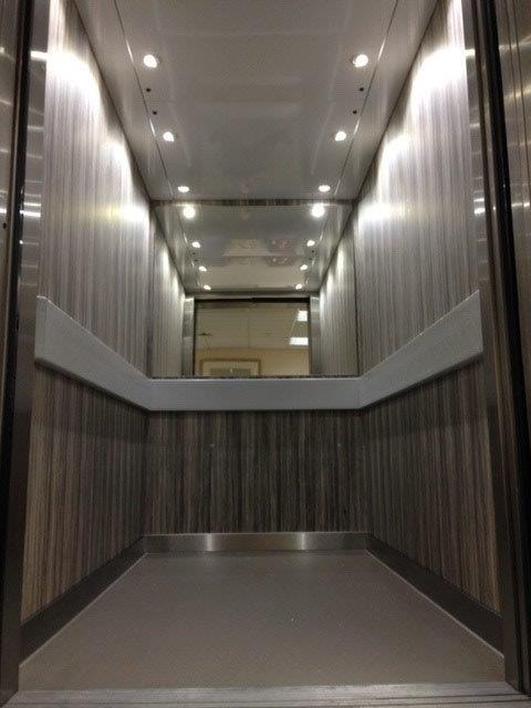 Interior of an elevator with wood-grain panels, overhead lighting, and a mirrored ceiling.