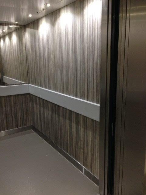 Elevator interior with wood-grain paneling, white handrail, and stainless steel door frame.