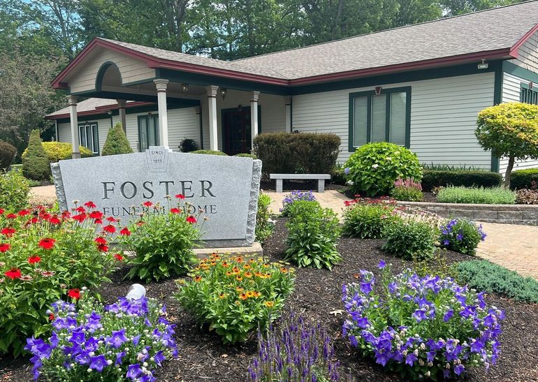 A gray stone sign for Foster Funeral Home sits in front of a white building surrounded by blooming red, purple, and green.