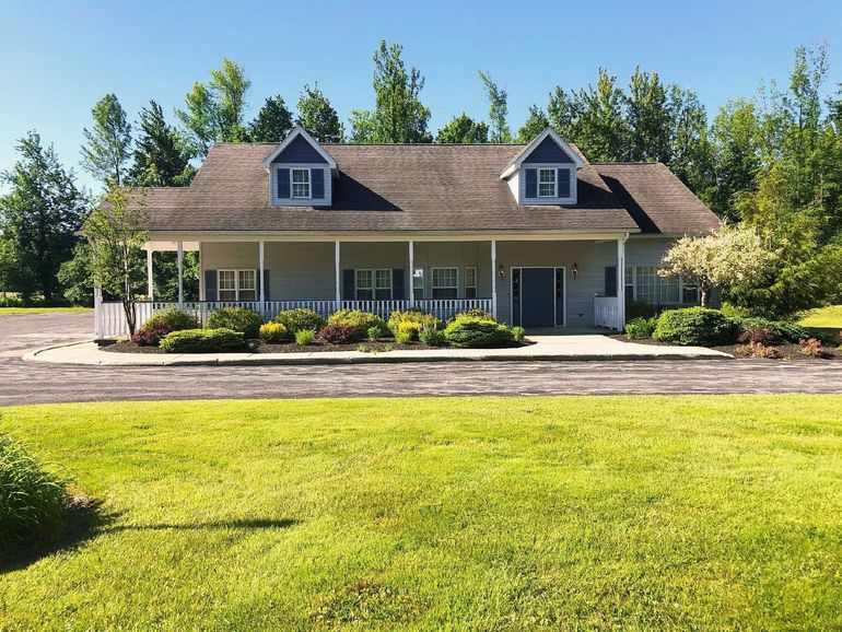 A gray farmhouse with a large front porch, dormer windows, and a brown roof, set against a green lawn and forest.