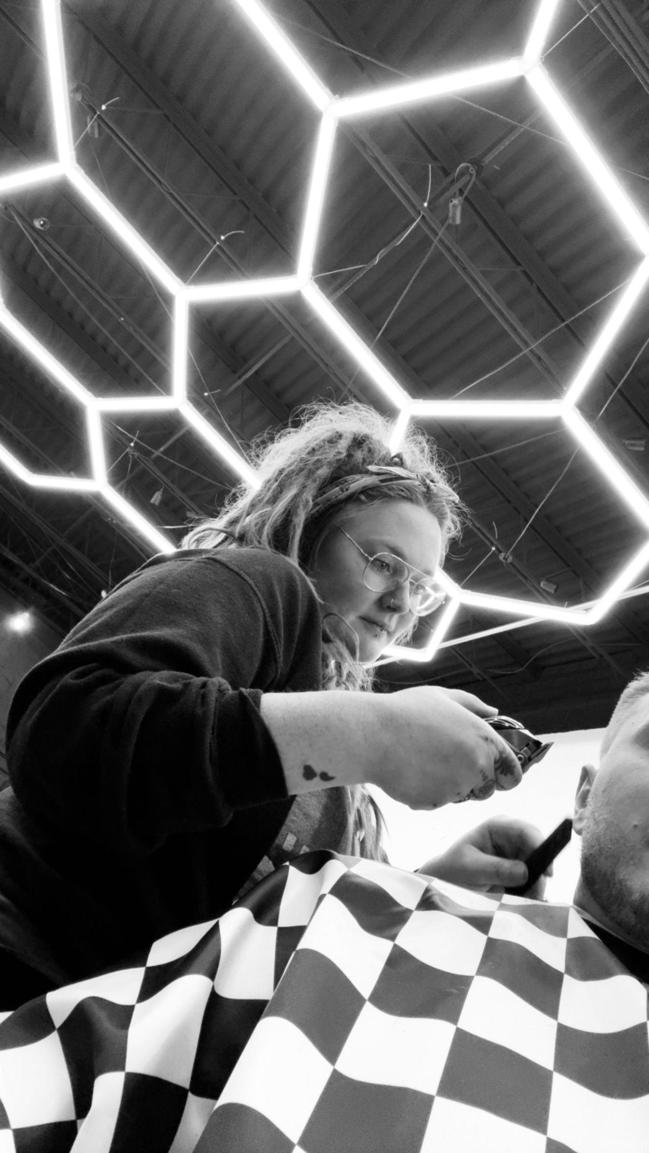 A hairstylist cuts hair in a salon with a bright, geometric hexagonal ceiling light. The client wears a checkered cape.