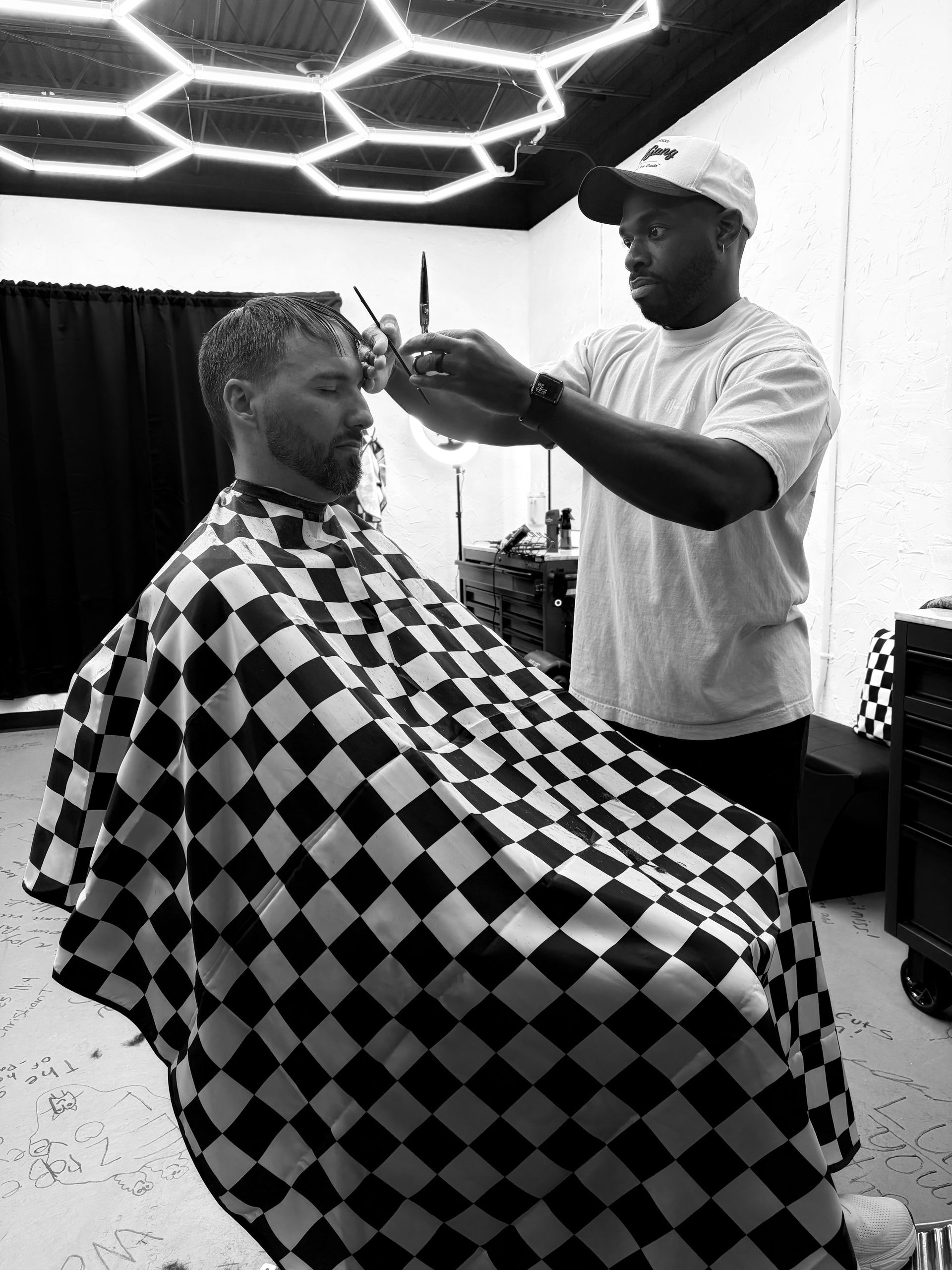 A barber trims a client’s hair in a studio with hexagonal ceiling lights, the client draped in a checkered cape.