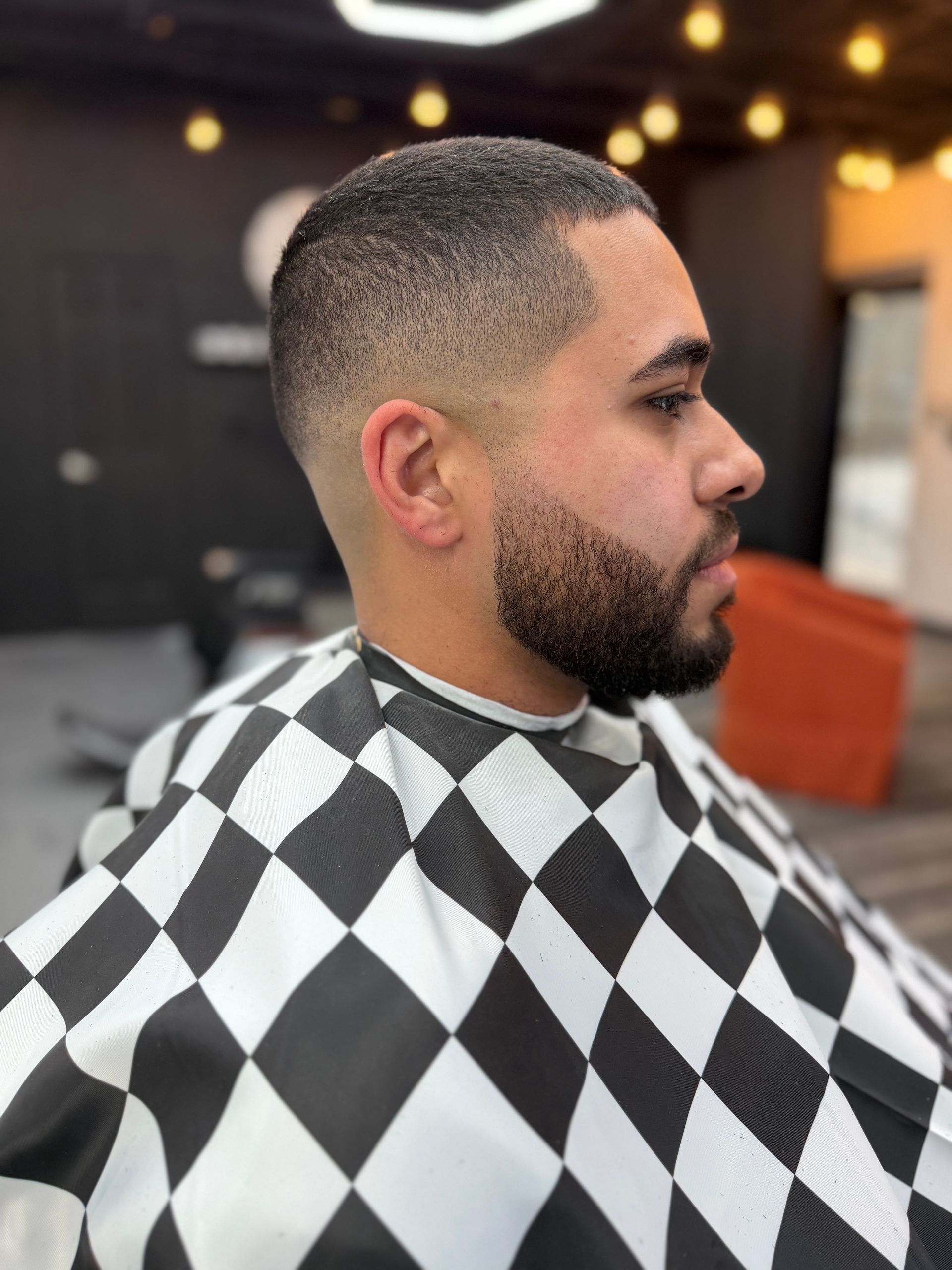 A person in a barber’s cape displays a short, precision-faded haircut and a neatly groomed beard in a salon setting.