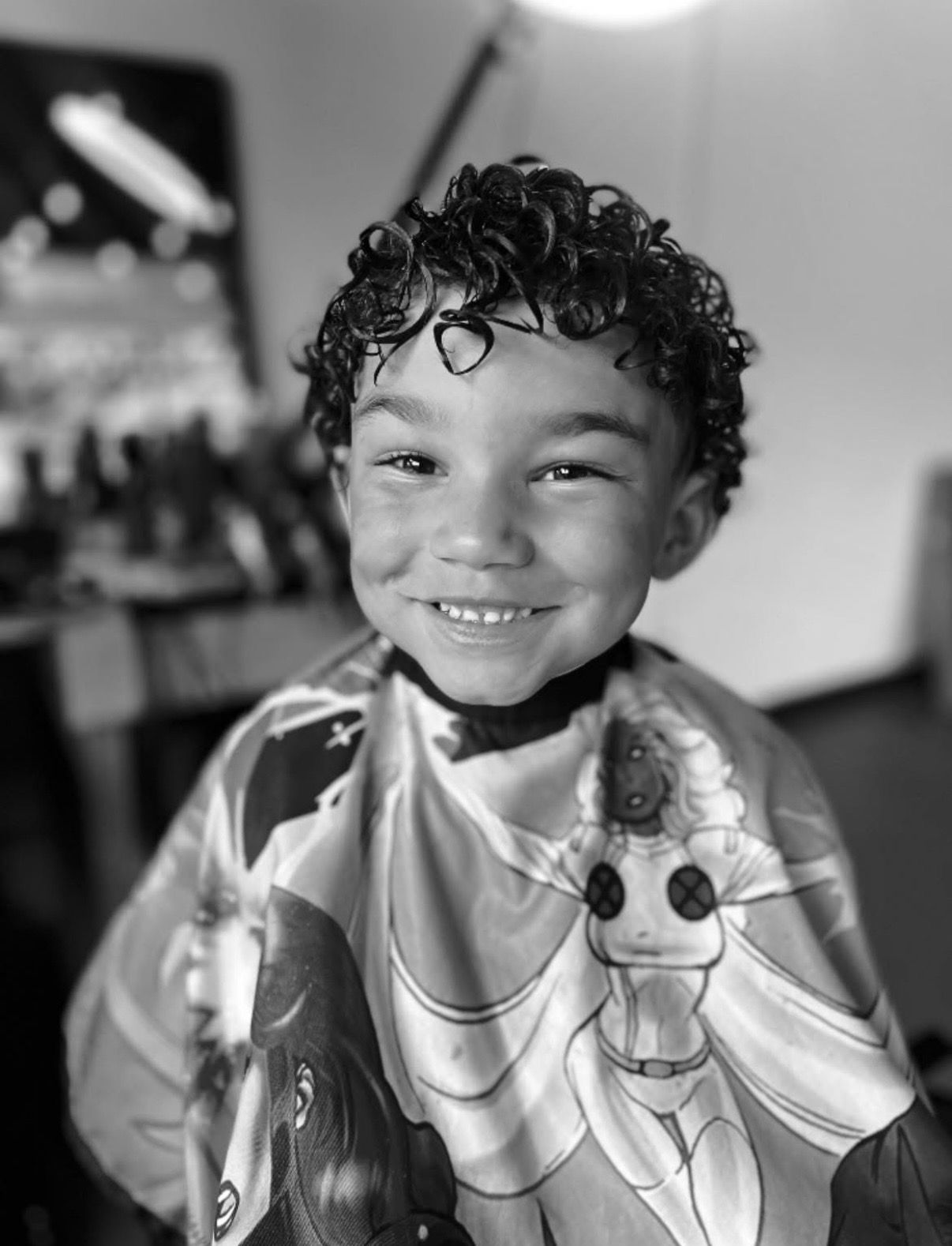 A smiling child wearing a barber cape with a graphic design sits in a salon chair.