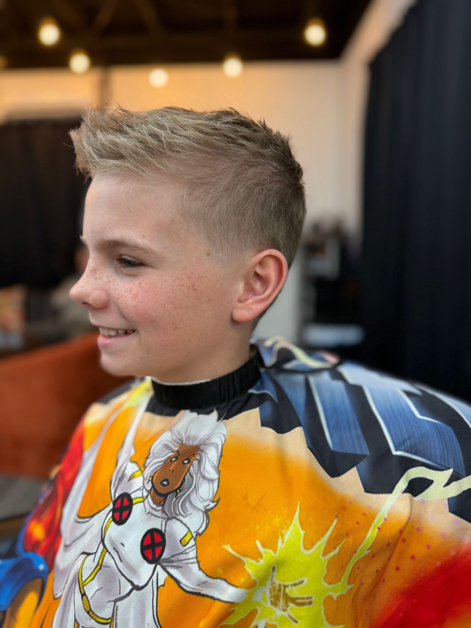 A person with a short, faded haircut and messy, textured hair on top, wearing a colorful X-Men-themed barber cape.