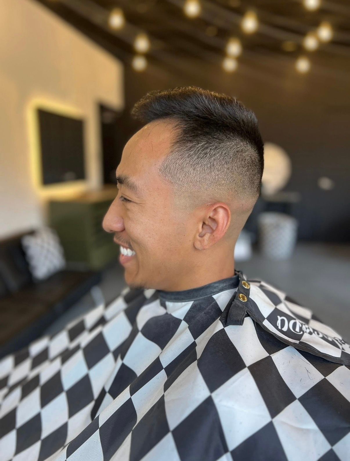 A smiling person sits in a barber's chair wearing a checkered cape, showcasing a fresh fade haircut in a salon.