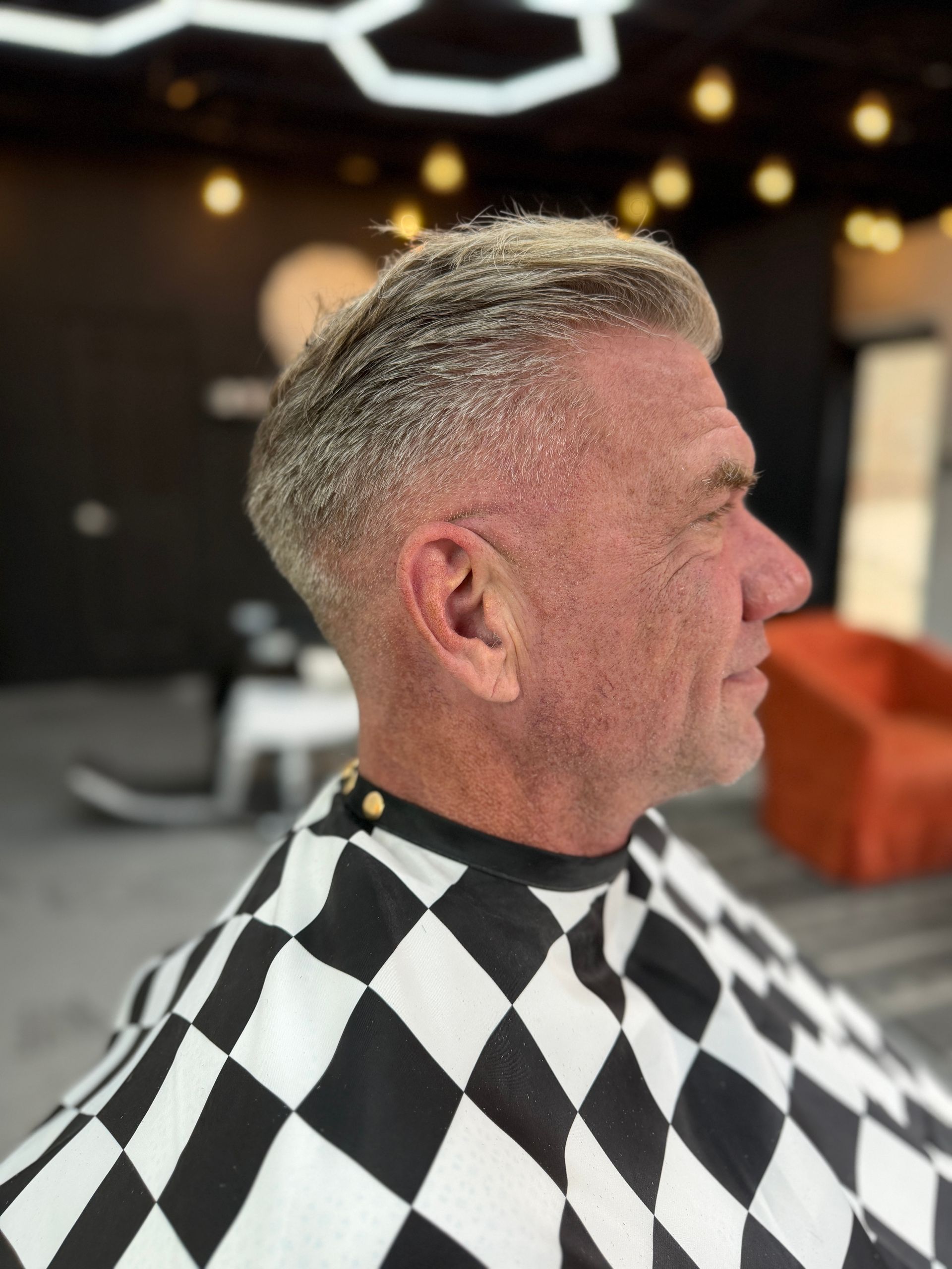 Side profile of a person in a barber's cape, featuring a short, textured, light-colored fade haircut in a salon.