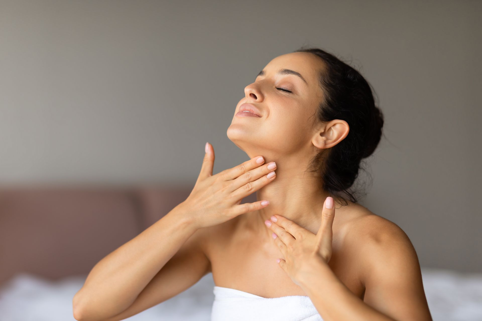 A woman wrapped in a towel is applying lotion to her neck.