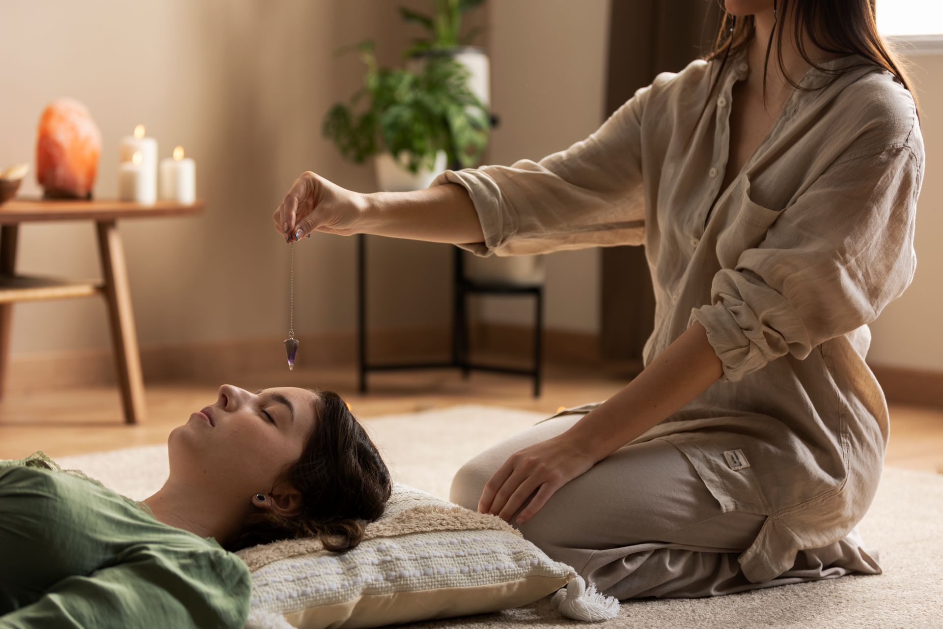 A woman is sitting on the floor holding a pendulum over a woman laying on the floor.