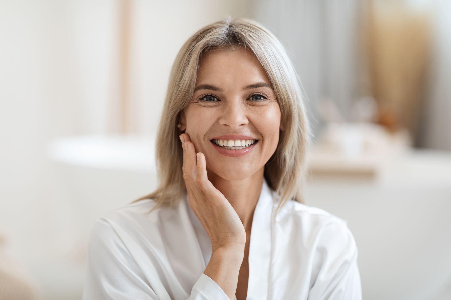 A woman in a white robe is smiling and touching her face.