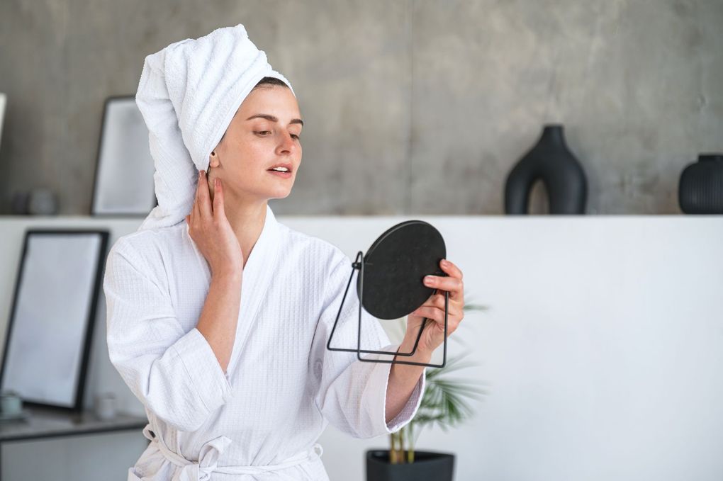 A woman with a towel wrapped around her head is looking at her face in a mirror.