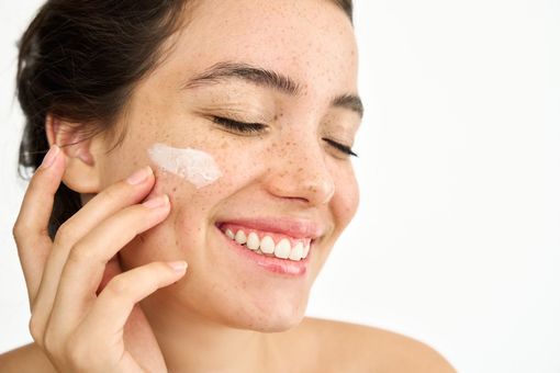 A woman is smiling while applying cream to her face.